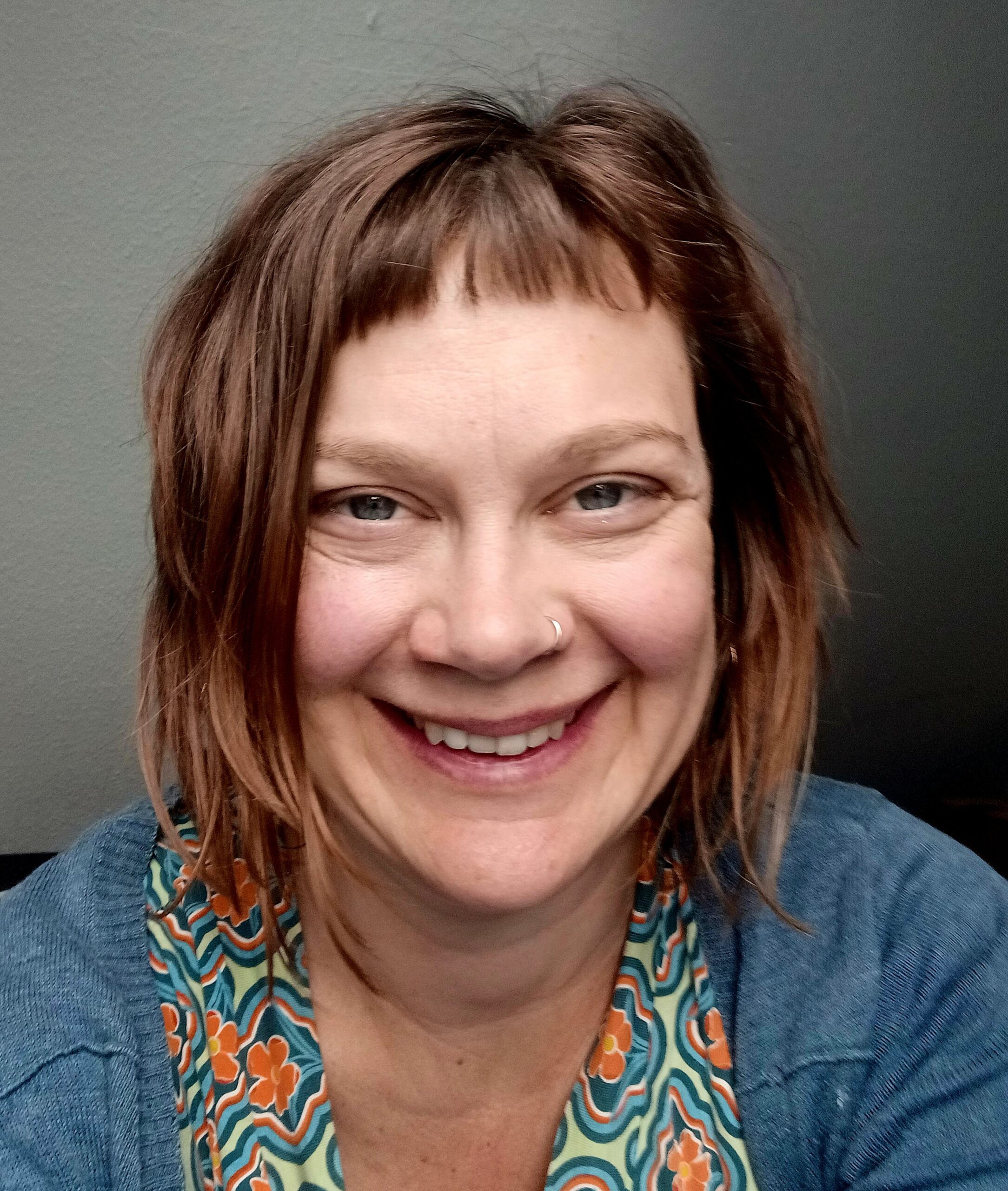 Portrait von Inga Schiffler. Sie trägt ihre Haare halblang und mit Pony, dazu ein buntes Shirt und eine blaue Jacke. Ihre Augen sind blau, die Nase klein und mit Nasenring. Sie lächelt.