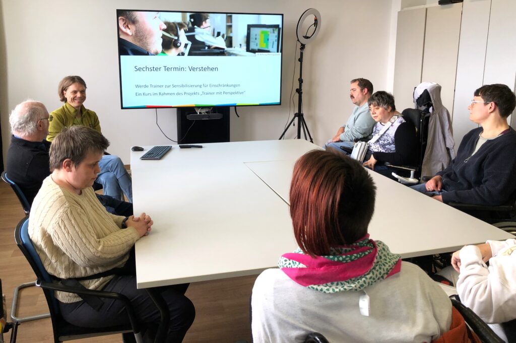 Menschen mit unterschiedlichen Behinderungen sitzen in einem Schulungsraum um einen großen Tisch. Ein Bildschirm in der Mitte zeigt eine Präsentationsfolie mit dem Titel: Sechster Termin: Verstehen.